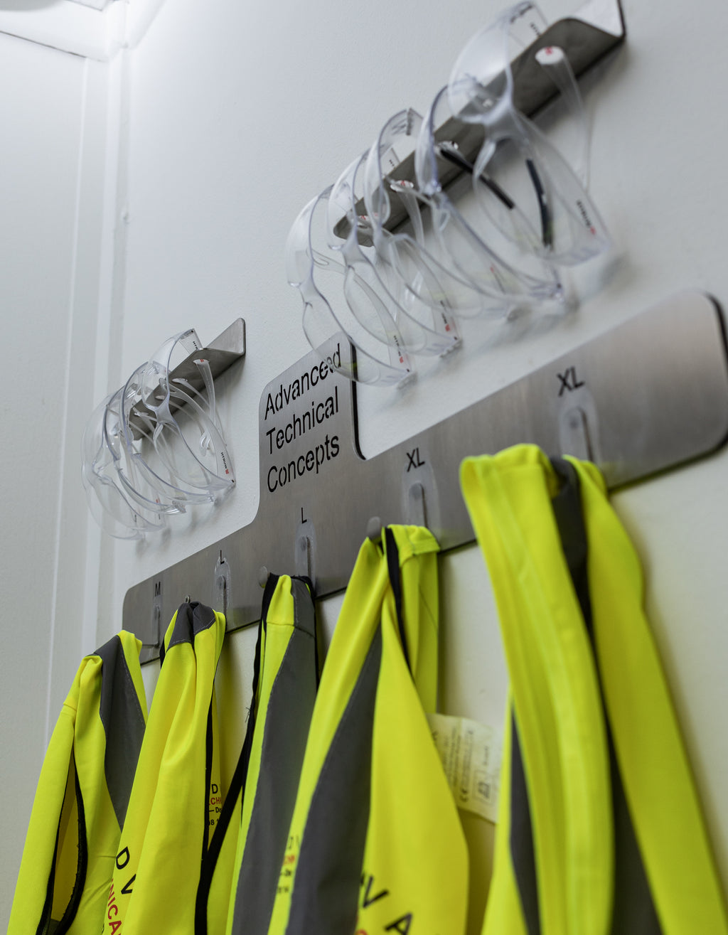 Hangers with safety vests on a wall labeled 'Advanced Technical Concepts'. The TLC Hi Viz Holder 5 Hangers keeps high visibility vests organized, accessible, and easily identifiable in Lean workplaces. It provides a dedicated storage solution for shared PPE in factories, warehouses, workshops, and site offices. This holder helps reduce clutter, prevent loss, and supports 5S and 6S safety standards. Ideal for entrances, locker areas, and workstations.