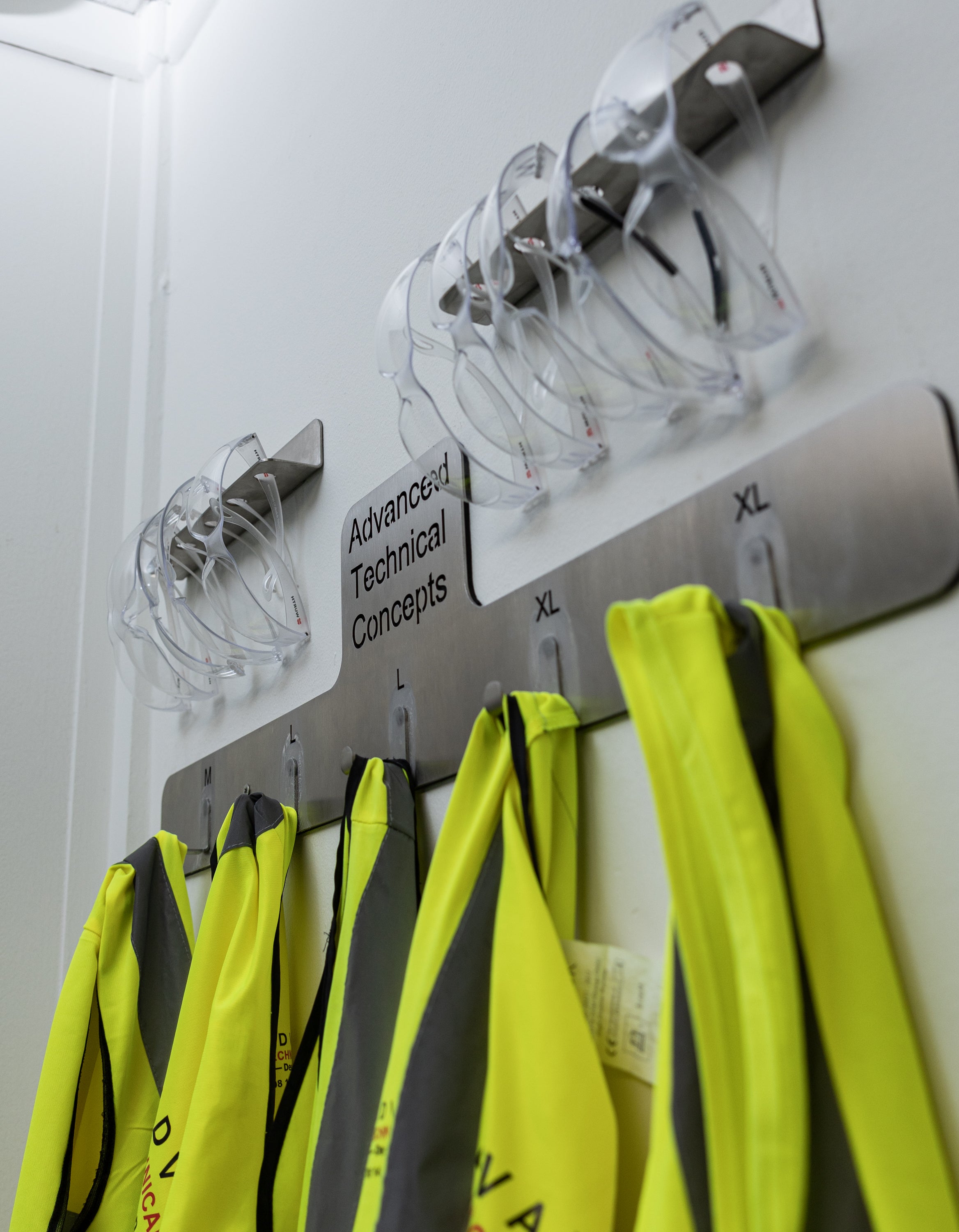 Hangers with safety vests on a wall labeled 'Advanced Technical Concepts'. The TLC Hi Viz Holder 5 Hangers keeps high visibility vests organized, accessible, and easily identifiable in Lean workplaces. It provides a dedicated storage solution for shared PPE in factories, warehouses, workshops, and site offices. This holder helps reduce clutter, prevent loss, and supports 5S and 6S safety standards. Ideal for entrances, locker areas, and workstations.